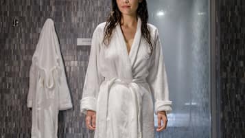 A person in a white robe standing in a modern bathroom with a tiled wall and a glass shower door.