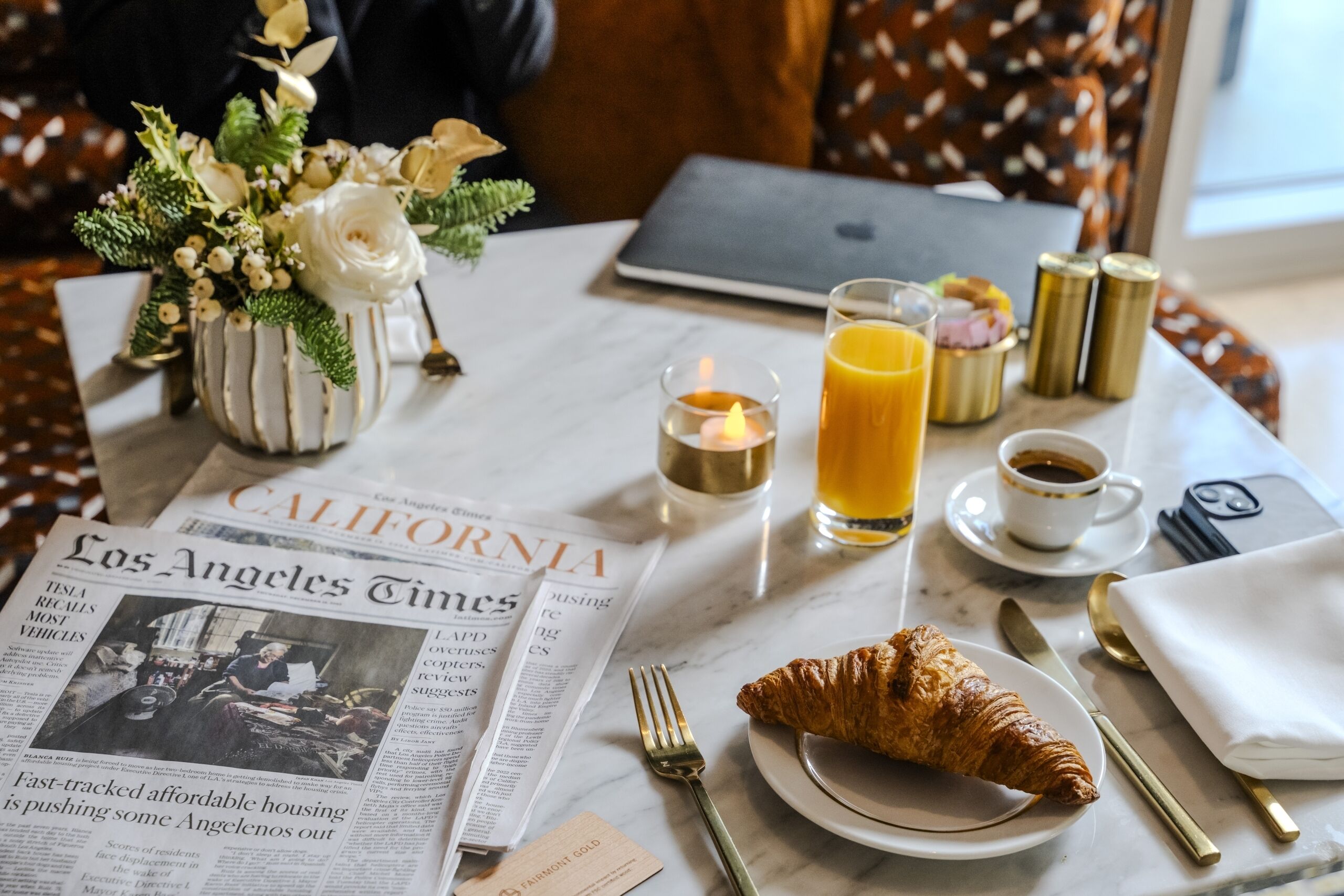 A breakfast setting with a newspaper, a croissant, a glass of orange juice, a cup of coffee, and a laptop on a table.
