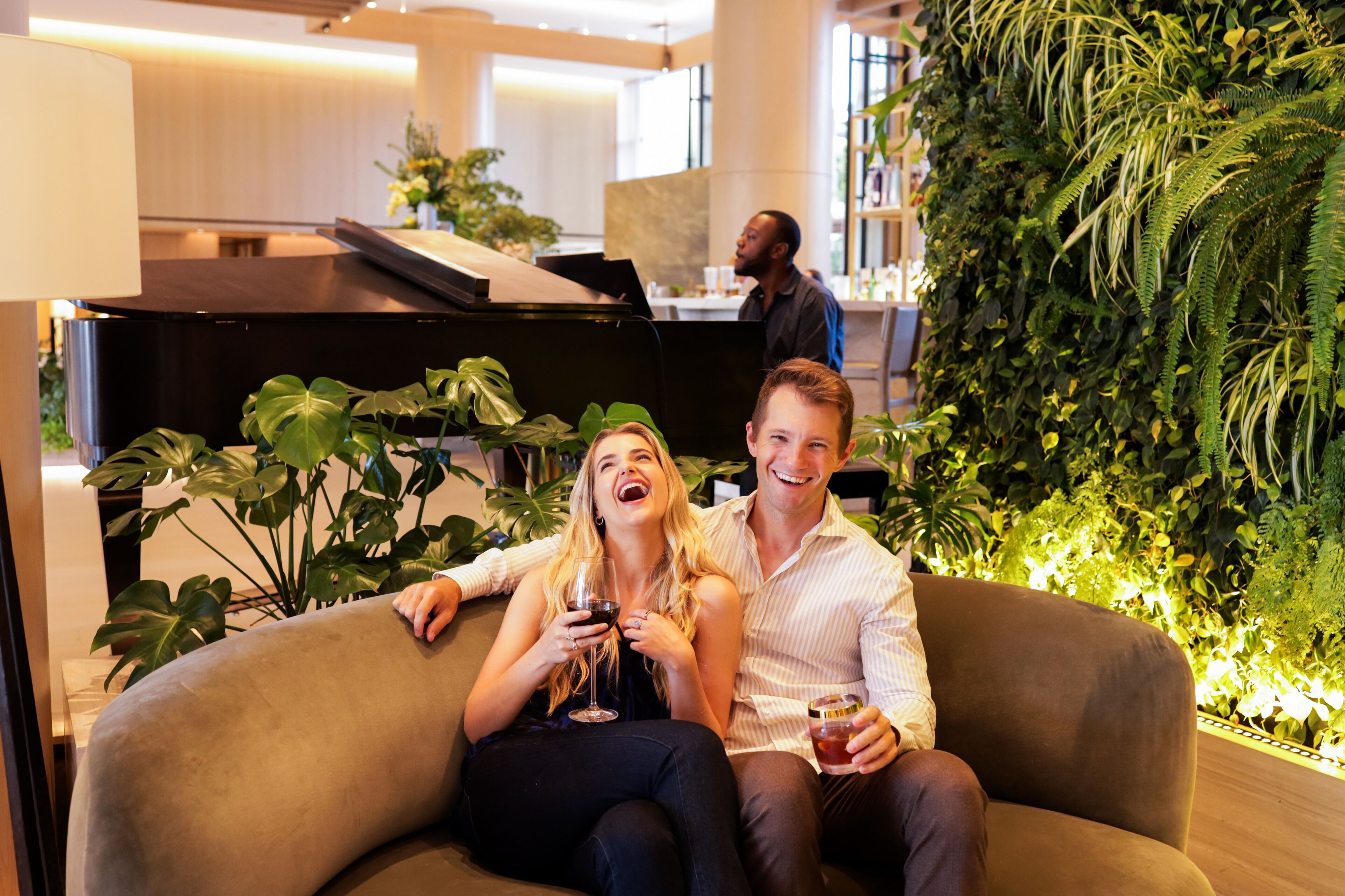 A couple sitting on a curved sofa in a modern indoor space with a green wall and a piano in the background.