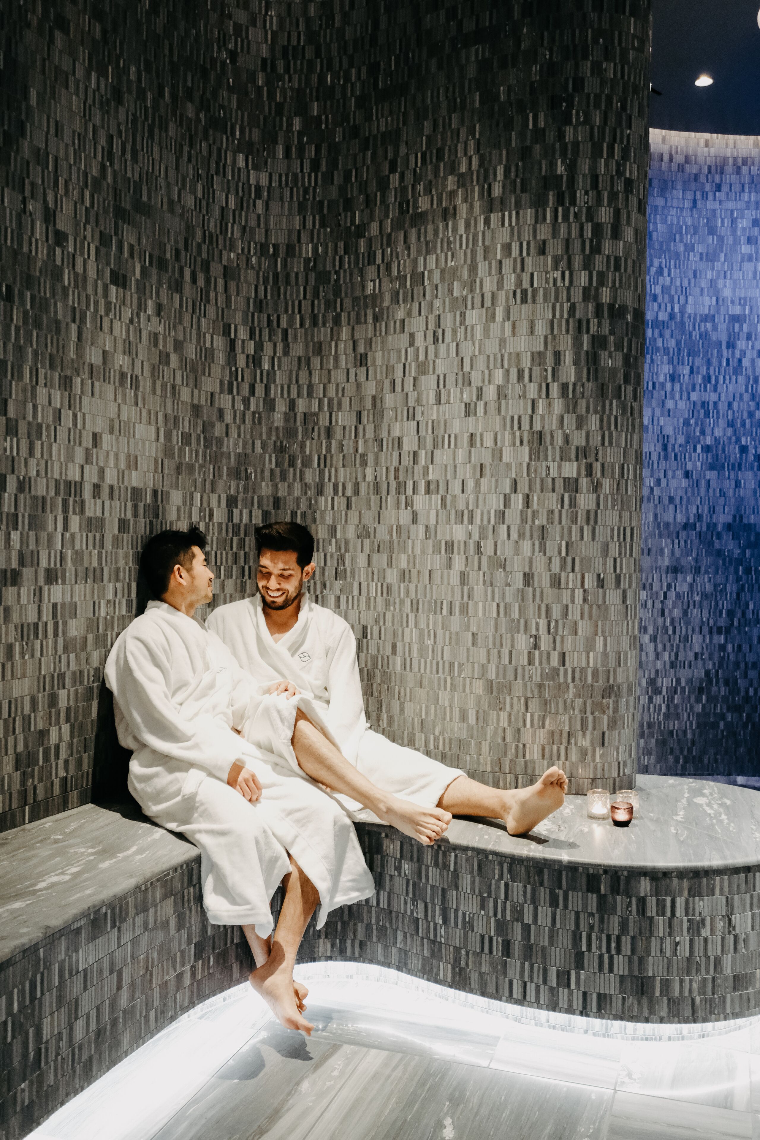 Two people in white robes sitting on a curved bench in a room with a textured wall and a glass floor.