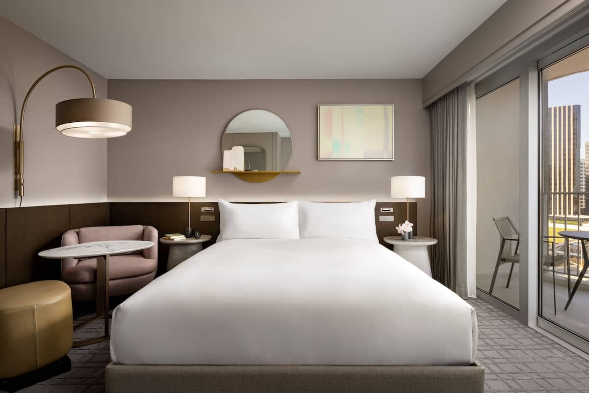 Ein modernes Hotelzimmer mit einem großen Bett, einem runden Spiegel, einem kleinen Tisch, einem Sessel und einem Balkon mit Blick auf die Stadt.