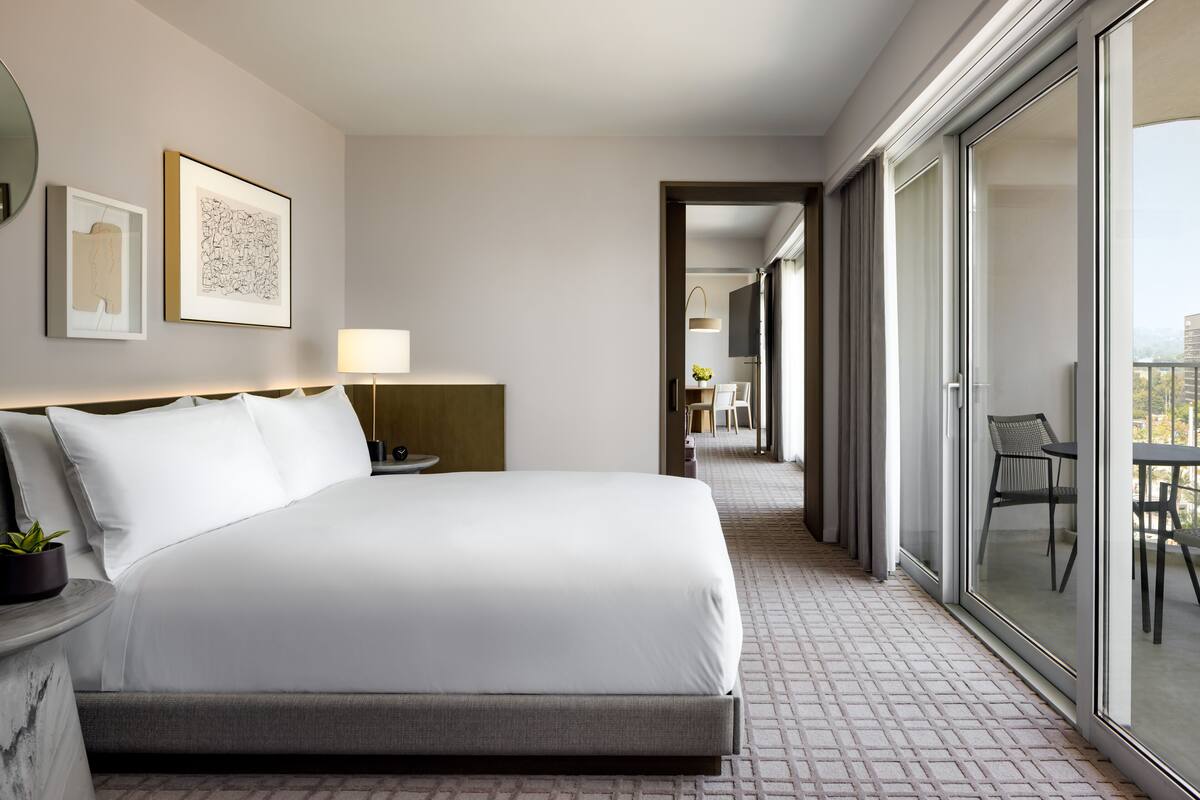 Ein modernes Hotelzimmer mit einem großen Bett, einem Nachttisch mit Lampe, einer kleinen Pflanze und Blick auf den Balkon durch die Glasdoor.