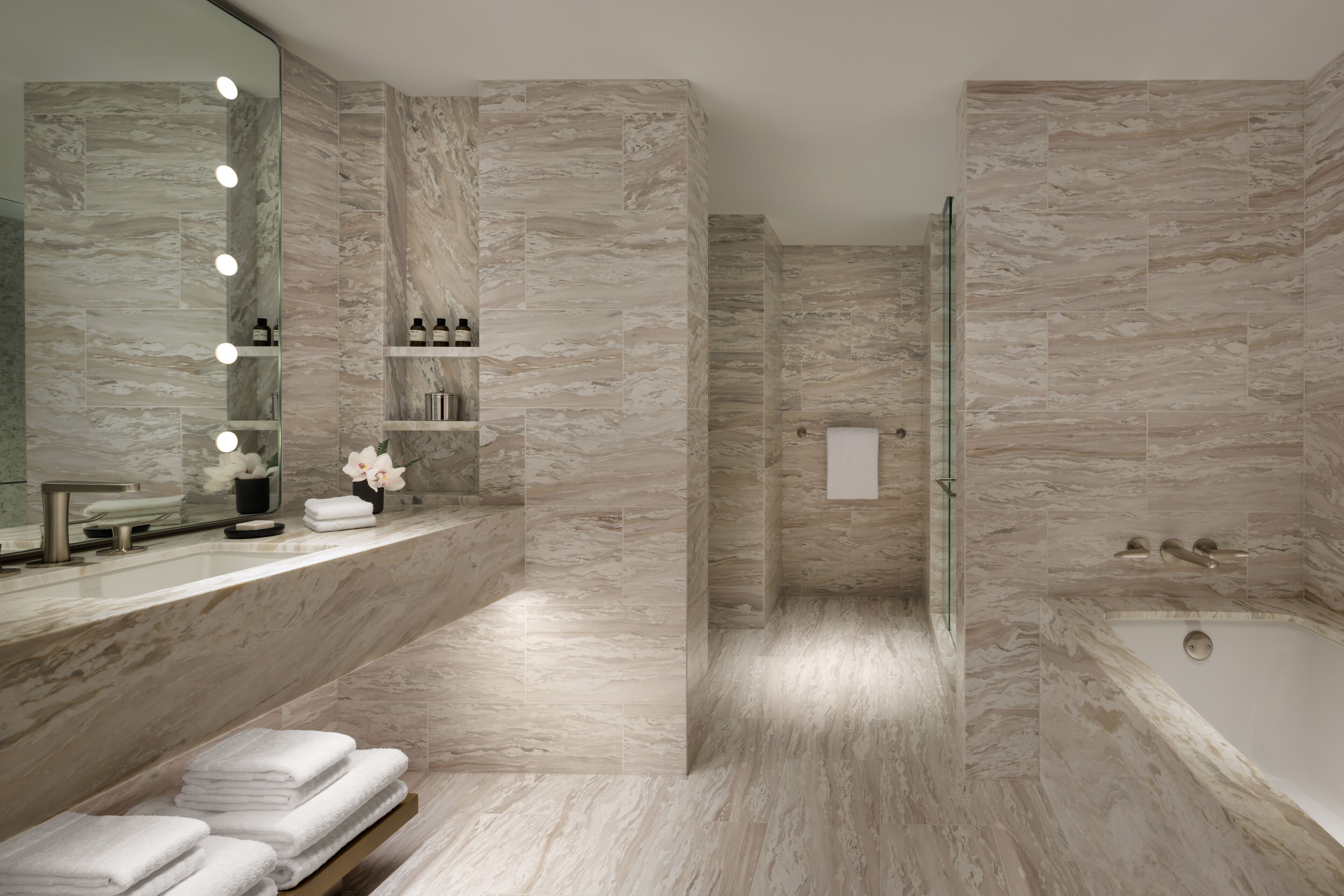 Un baño moderno con pared de mármol, cabina de ducha de vidrio, tocador con espejo y bañera.