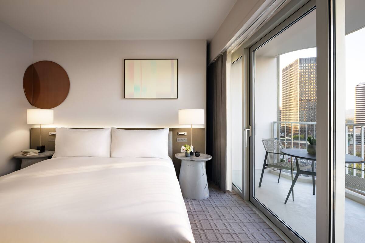 Ein Hotelzimmer mit einem großen Bett, zwei Nachttischlampen, einer runden Wanddekoration, einem gerahmten abstrakten Gemälde und einem Balkon mit Tisch und Stühlen.