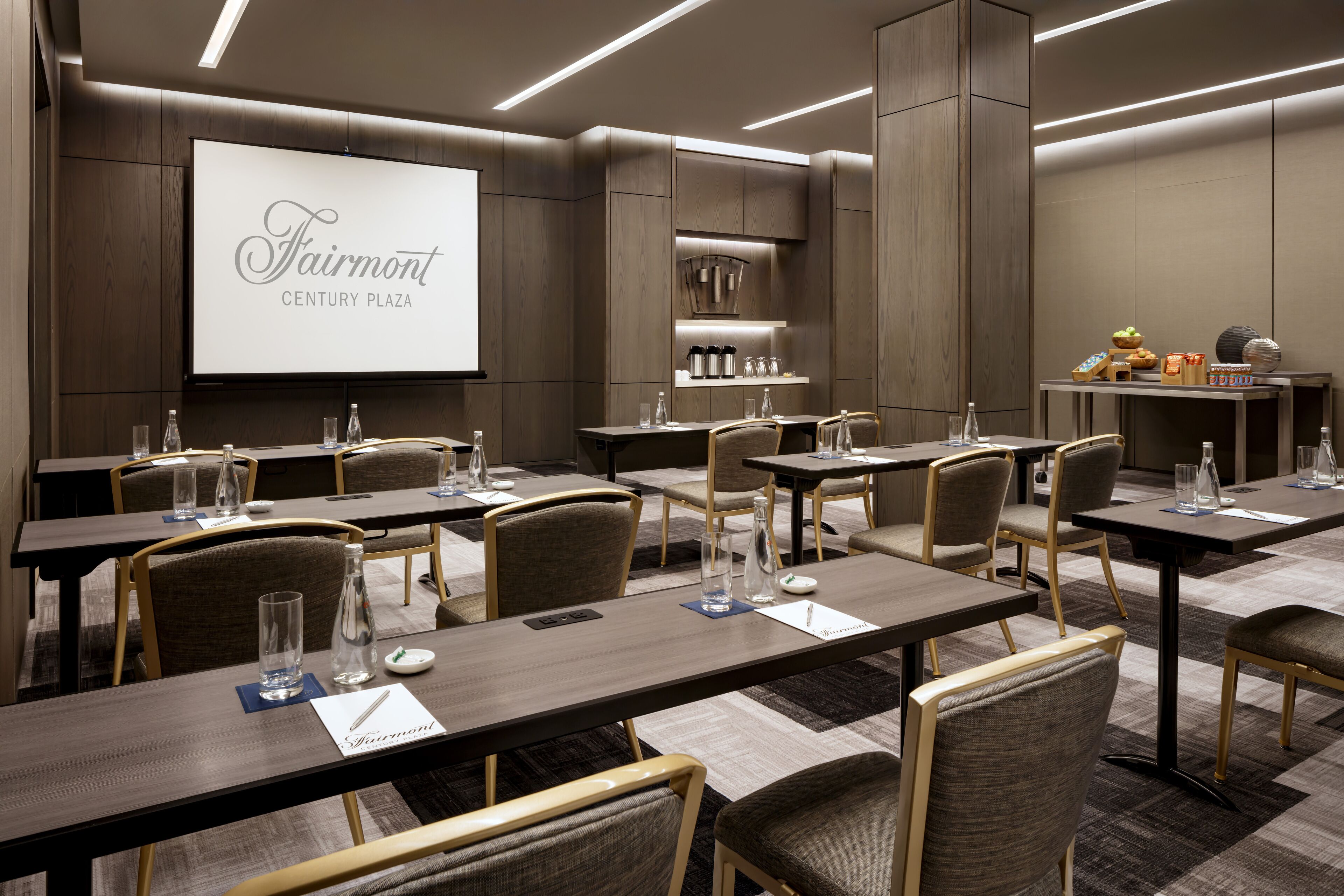 A conference room with multiple tables and chairs, a large screen displaying "Fairmont CENTURY PLAZA," and a small kitchenette area.