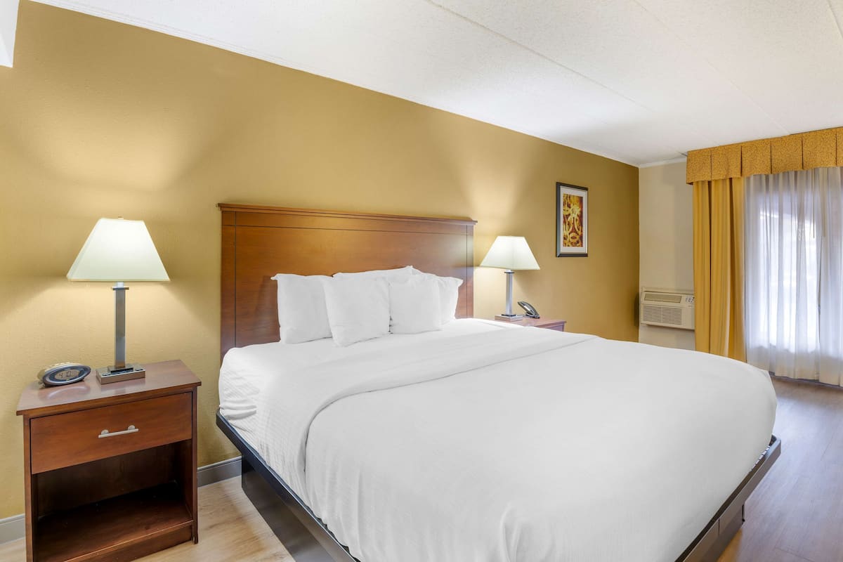 A hotel room with a large bed, two bedside tables with lamps, a wooden nightstand, a wall-mounted phone, and a window with curtains.