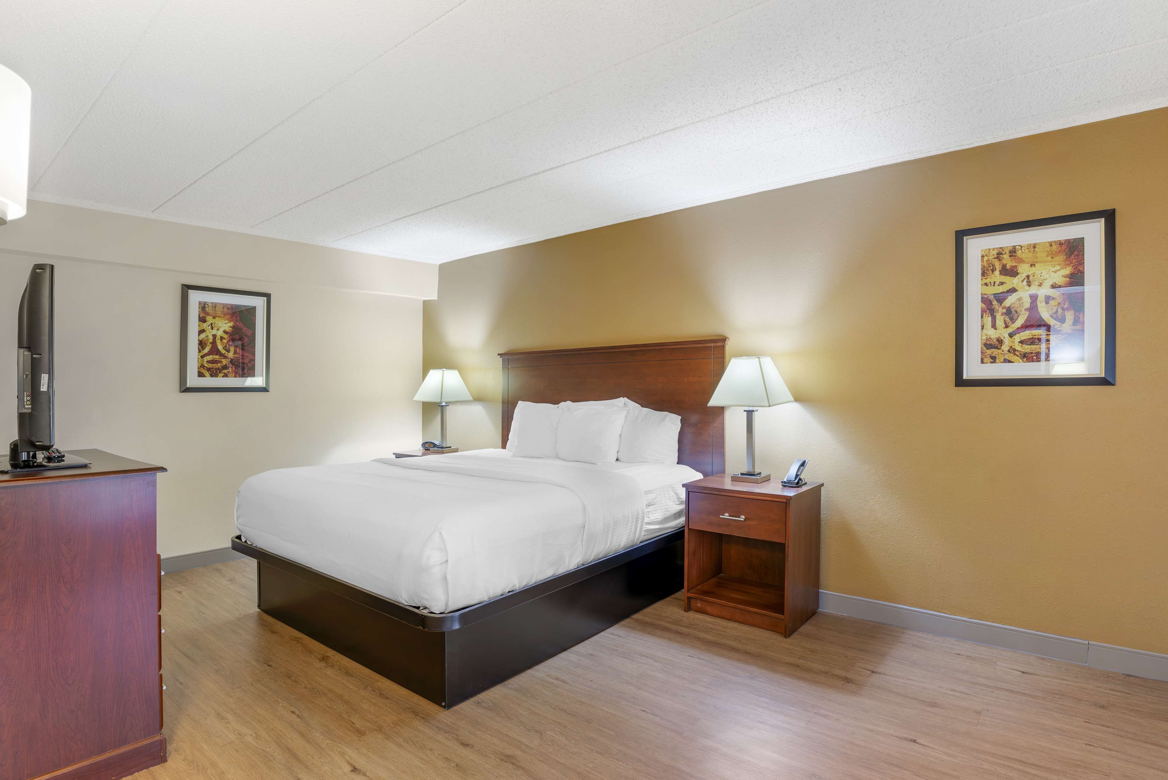A hotel room with a bed, bedside lamps, a wooden nightstand, and artwork on the walls.