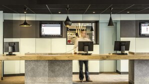 A modern reception desk with a wooden counter, two monitors, and pendant lighting. - ibis Paris Porte de Bagnolet (Bagnolet)