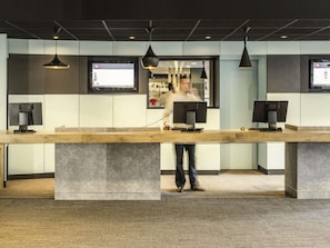 A modern reception desk with a wooden counter, two monitors, and pendant lighting. - ibis Paris Porte de Bagnolet (Bagnolet)