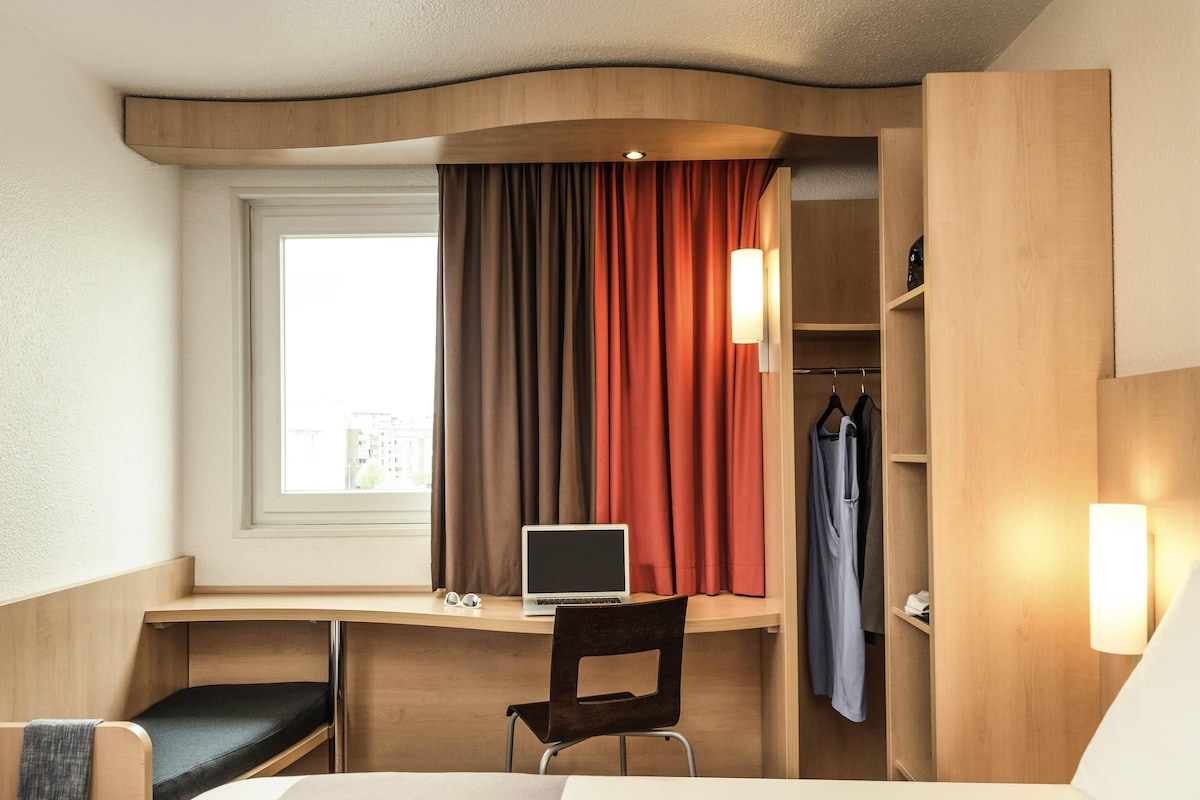 A hotel room with a bed, desk, chair, laptop, window, curtains, and a wardrobe.