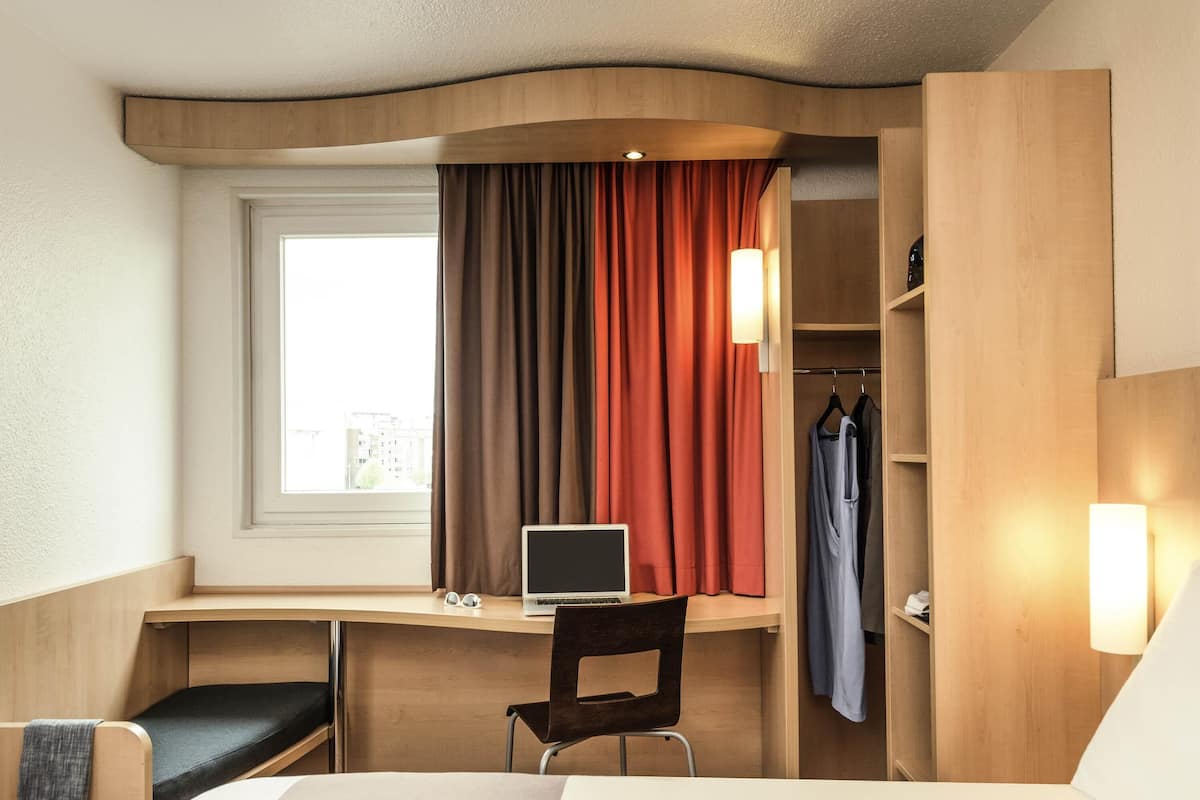 Une chambre d’hôtel équipée d’un lit, d’un bureau, d’une chaise, d’un ordinateur portable, d’une fenêtre, de rideaux et d’une armoire.