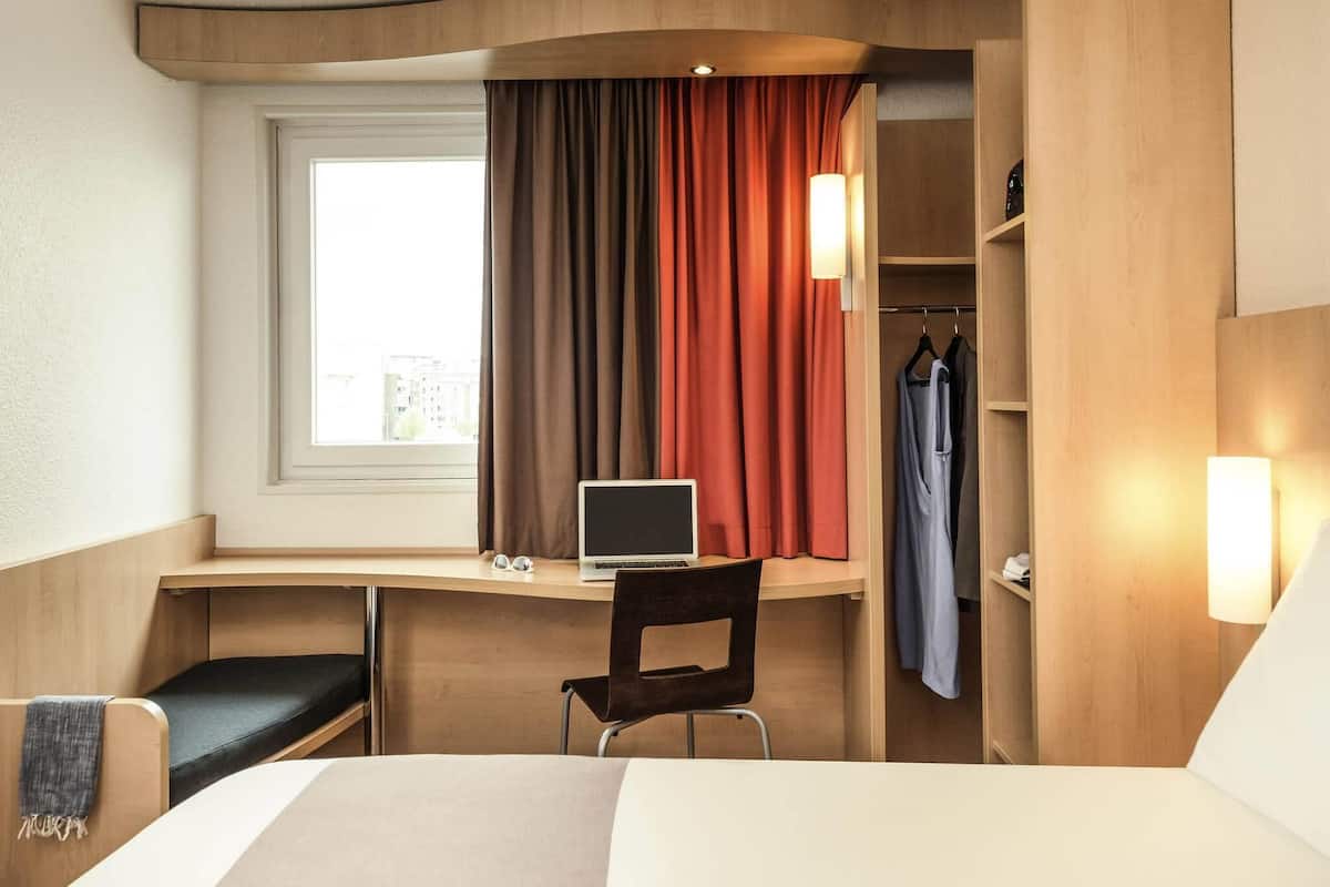 A hotel room with a desk, a laptop, a chair, a bed, a window with curtains, and a wardrobe.