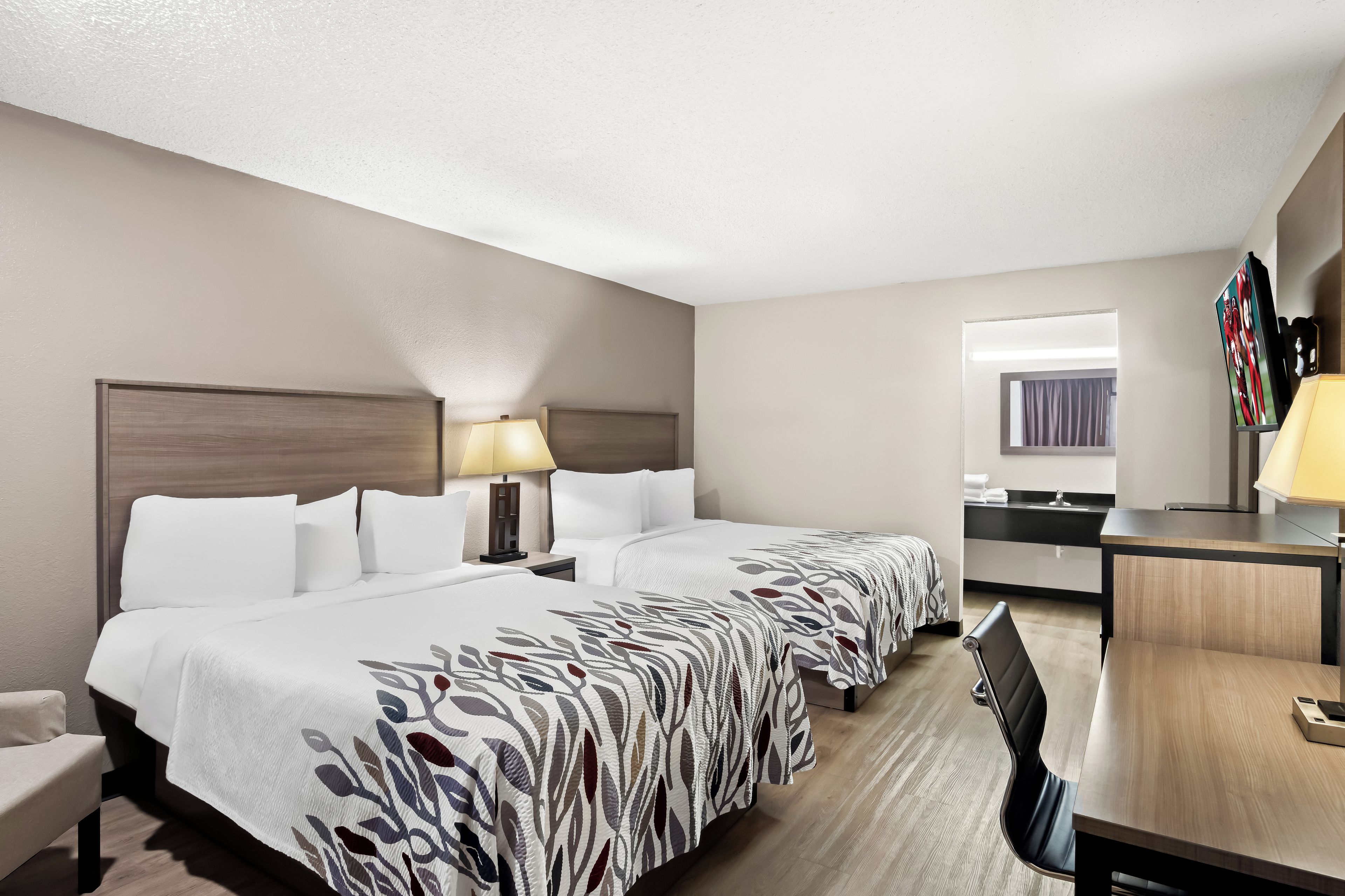 A hotel room with two beds, a desk, a chair, a lamp, and a television.