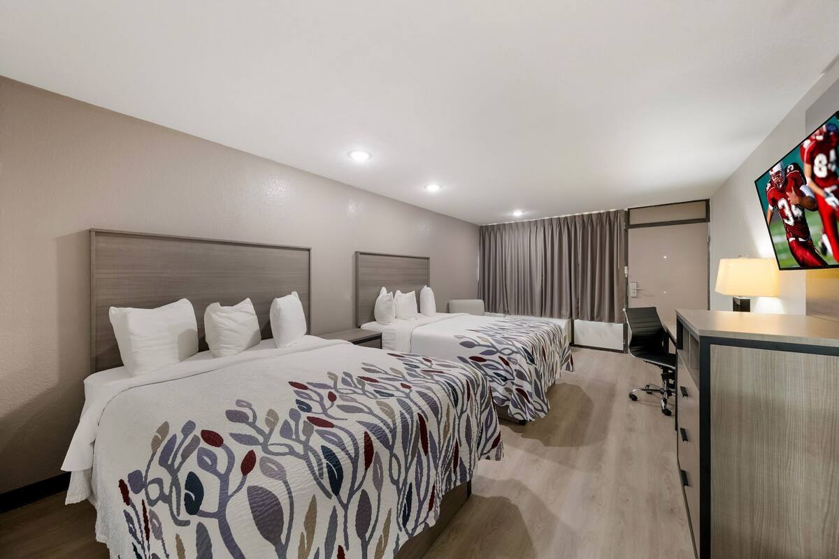 A hotel room with three beds, a desk, a TV, and a lamp.