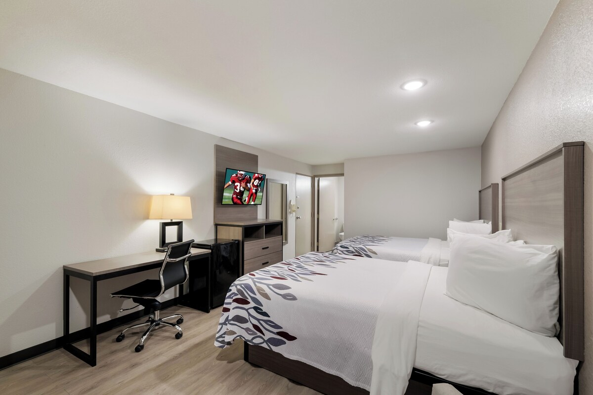 A hotel room with a desk, chair, two beds, a TV, and a lamp.