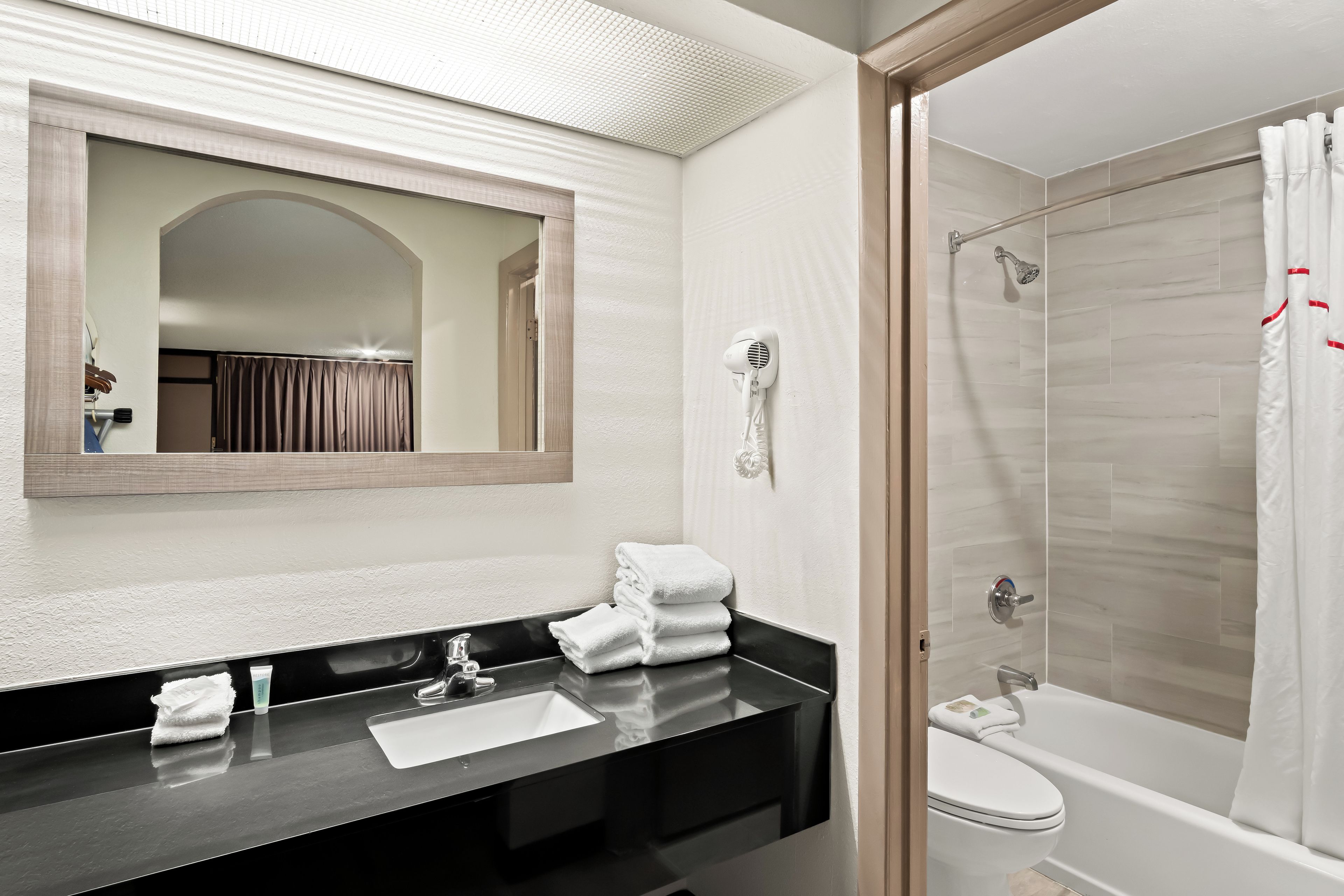 A bathroom with a black countertop, a mirror, a hairdryer, a sink, and a shower area.