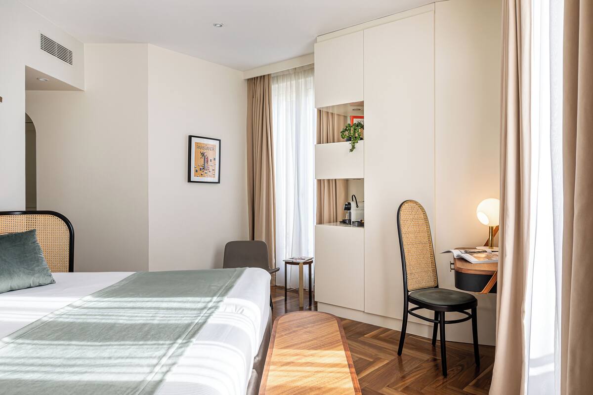 A hotel room with a bed, a desk, a chair, a small table, a potted plant, and a wall-mounted shelf.