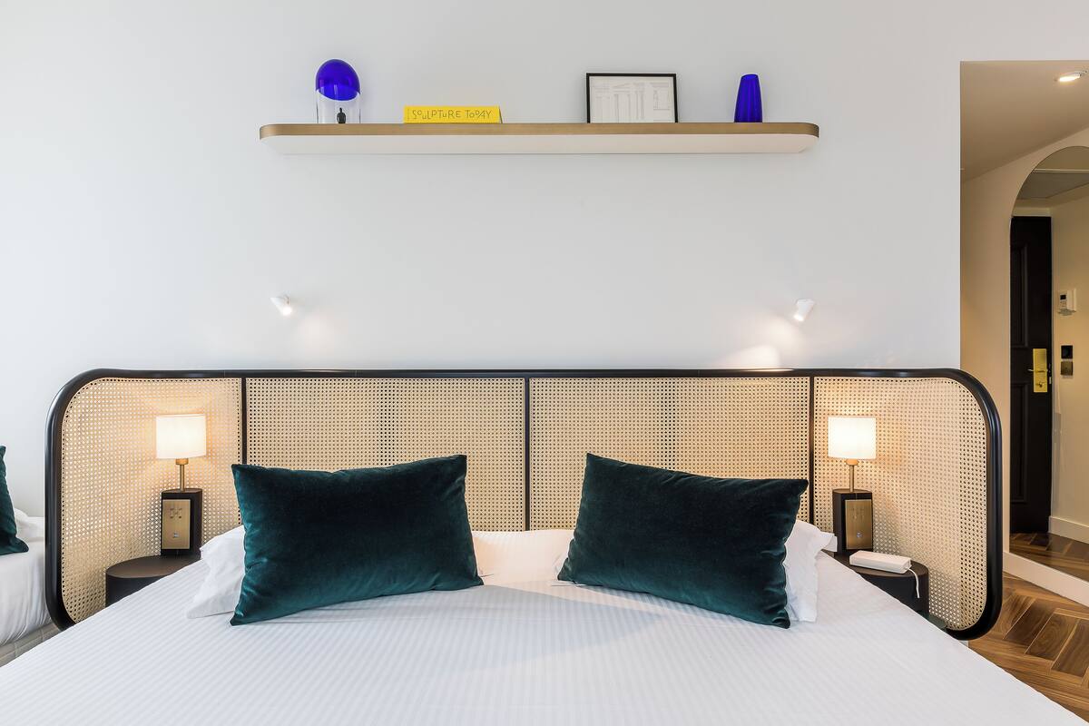 A hotel room with a bed, two bedside lamps, a shelf with decorative items, and a door leading to another room.