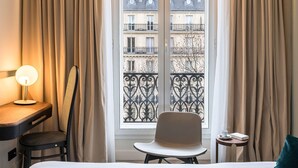 A hotel room with a bed, a desk with a lamp, a chair, a small table with a book and a cup, and a view of buildings through a window with curtains. - Hotel le Friedland (Paris)