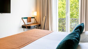 A hotel room with a bed, a desk with a lamp, and a view of greenery outside. - Hotel le Friedland (Paris)