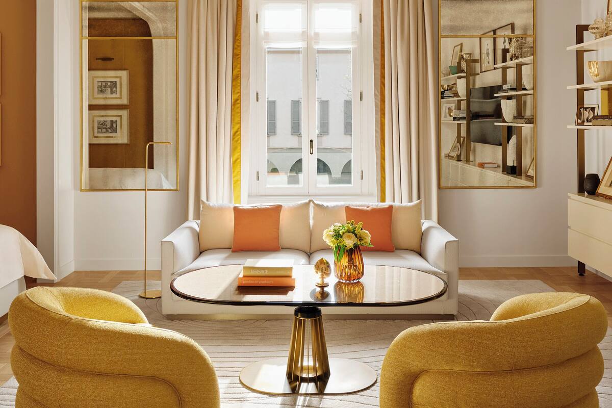 A room with a white sofa, yellow armchairs, a round coffee table, a large mirror, and a ceiling with intricate designs.