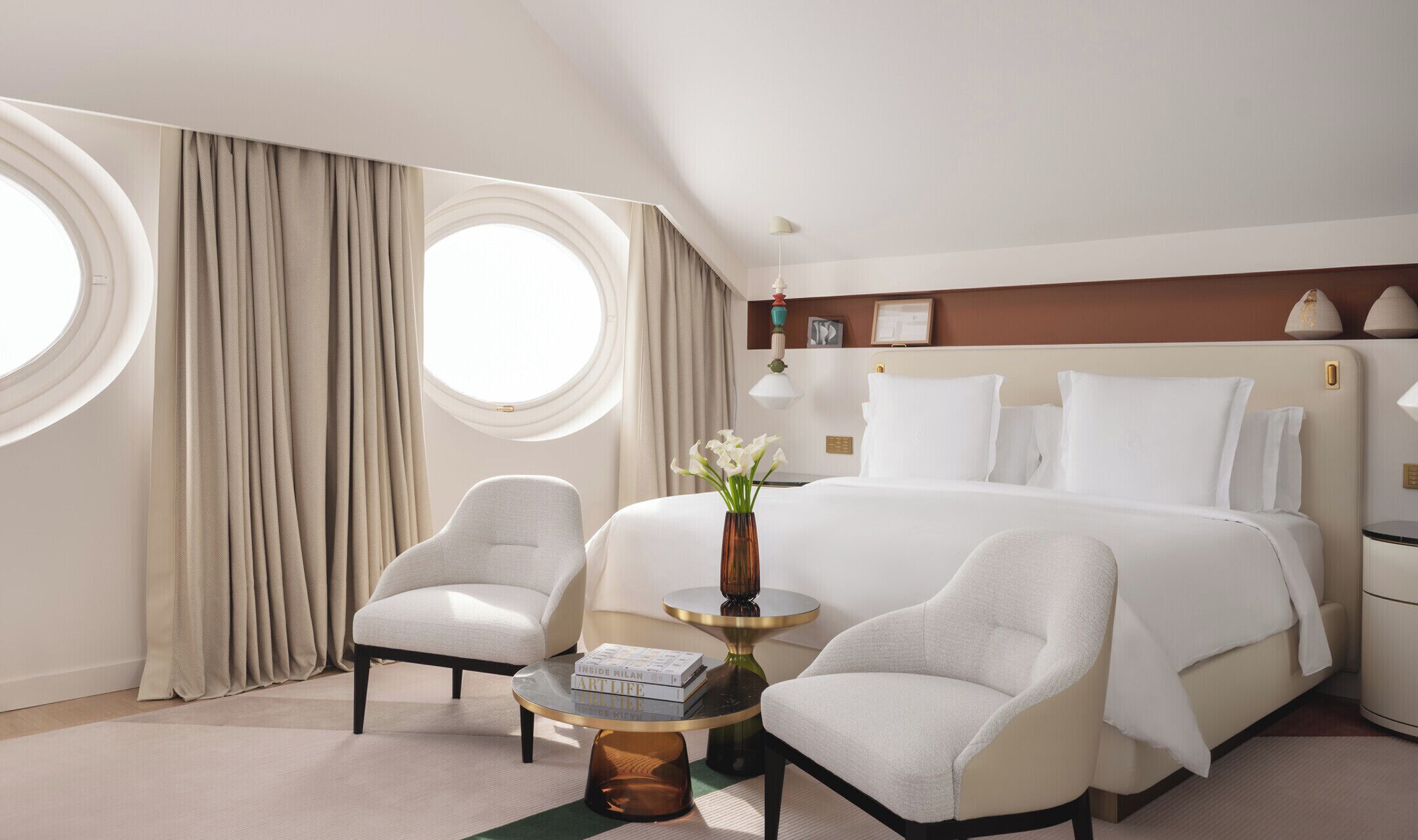 Una habitación de hotel con una cama grande, dos sillones, una mesita redonda con un florero y una ventana con cortinas.