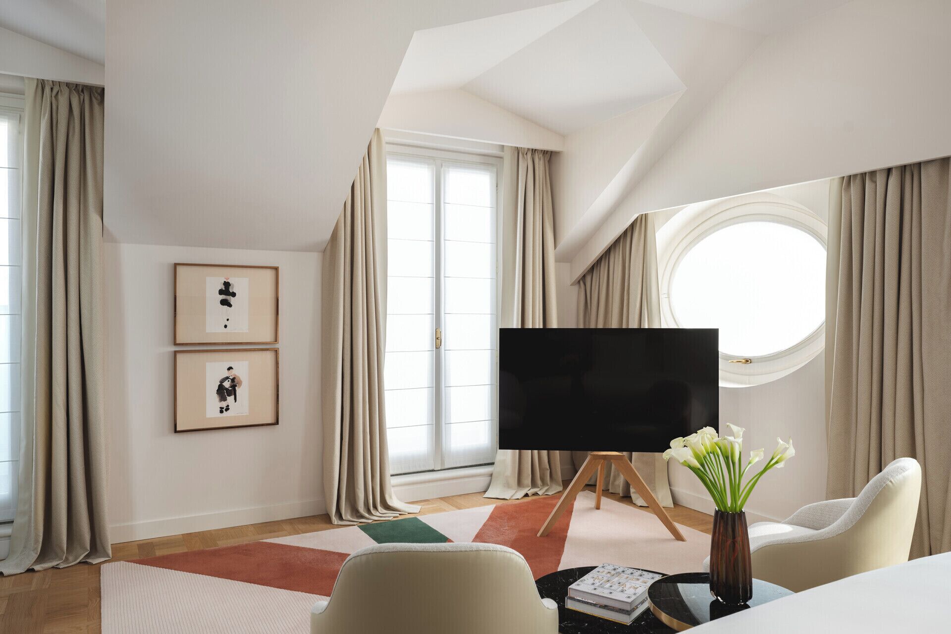 A modern hotel room with a large window, a flat-screen TV, a round window, and a vase with flowers.