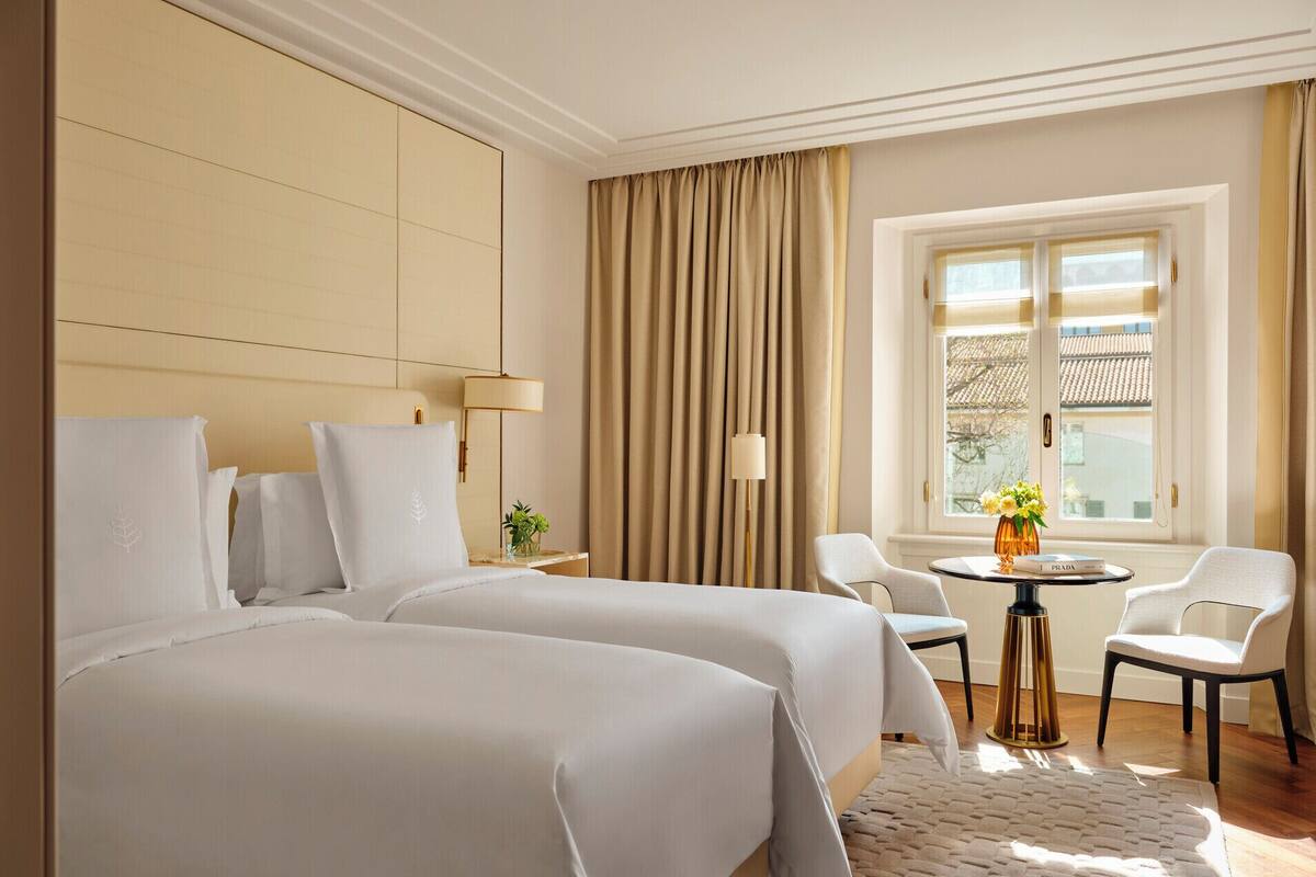 A hotel room with two beds, a small table with chairs, a window with curtains, and a vase with flowers.