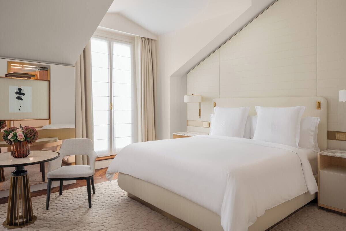 A hotel room with a large bed, a desk with a chair, a bedside table, and a large window with curtains.