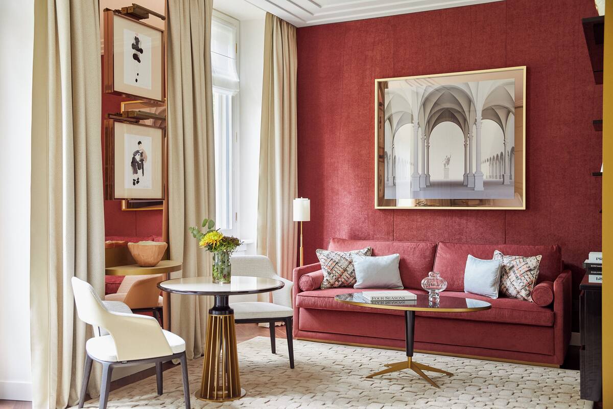 A room with a red wall, a large framed artwork, a red sofa with patterned cushions, a round coffee table, and a white chair.