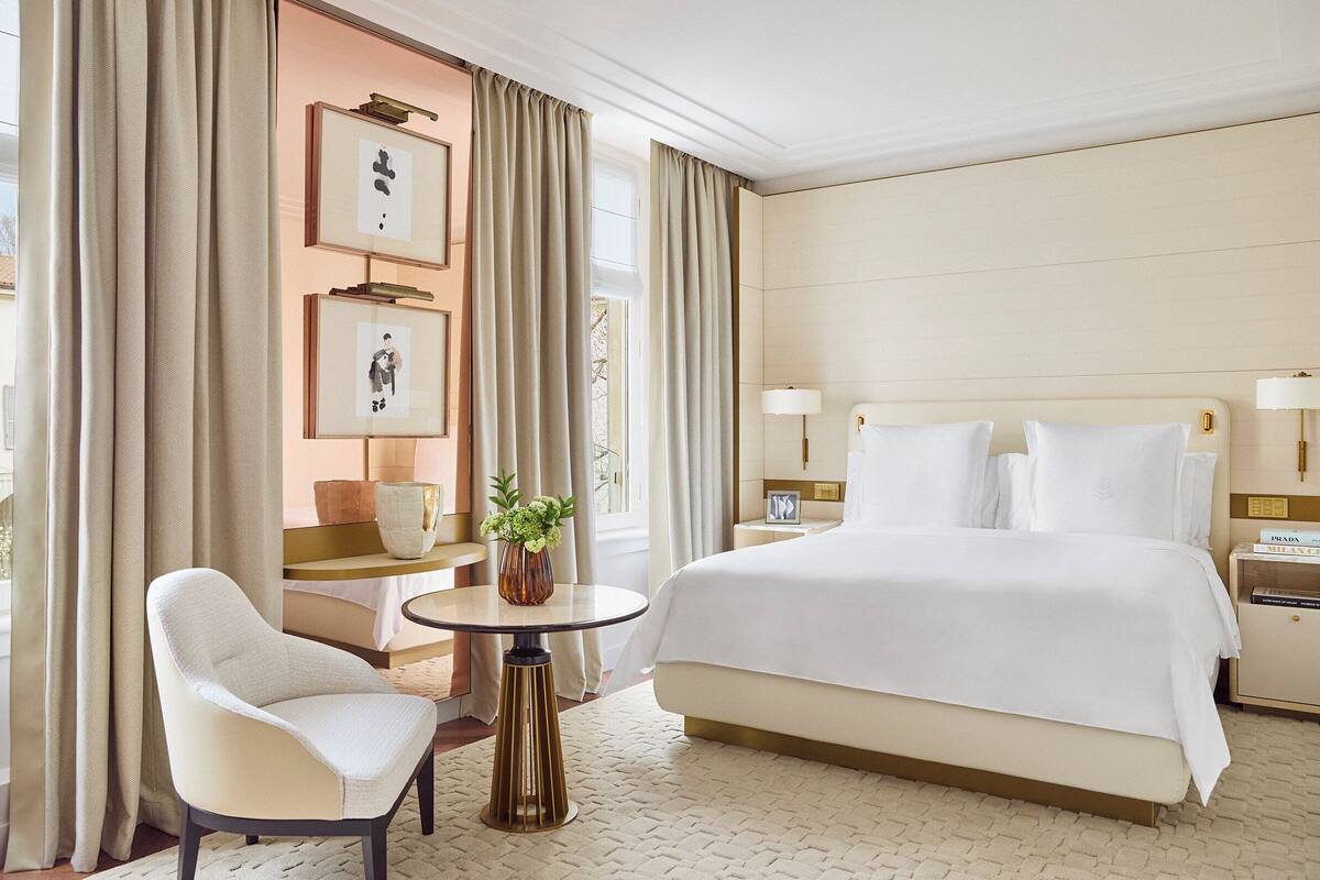 A hotel room with a large bed, a chair, a small table with a vase, and a nightstand with a lamp.
