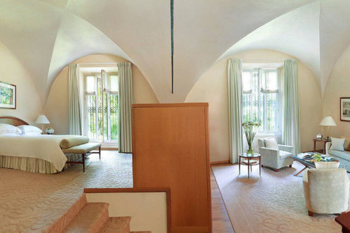 A spacious hotel room with a large bed, two seating areas, and a view of the outdoors through arched windows.