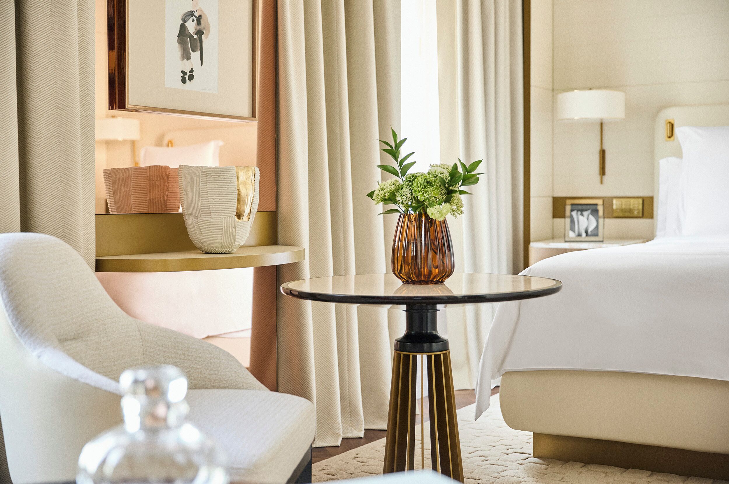 A hotel room with a bed, a chair, a small round table with a vase of flowers, and a nightstand with a lamp.