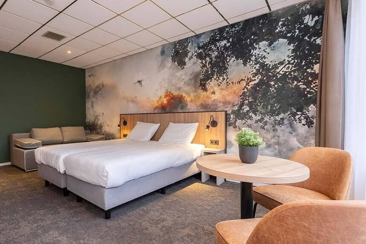 A hotel room with a large bed, a bedside table, a chair, a small round table, and a wall mural.
