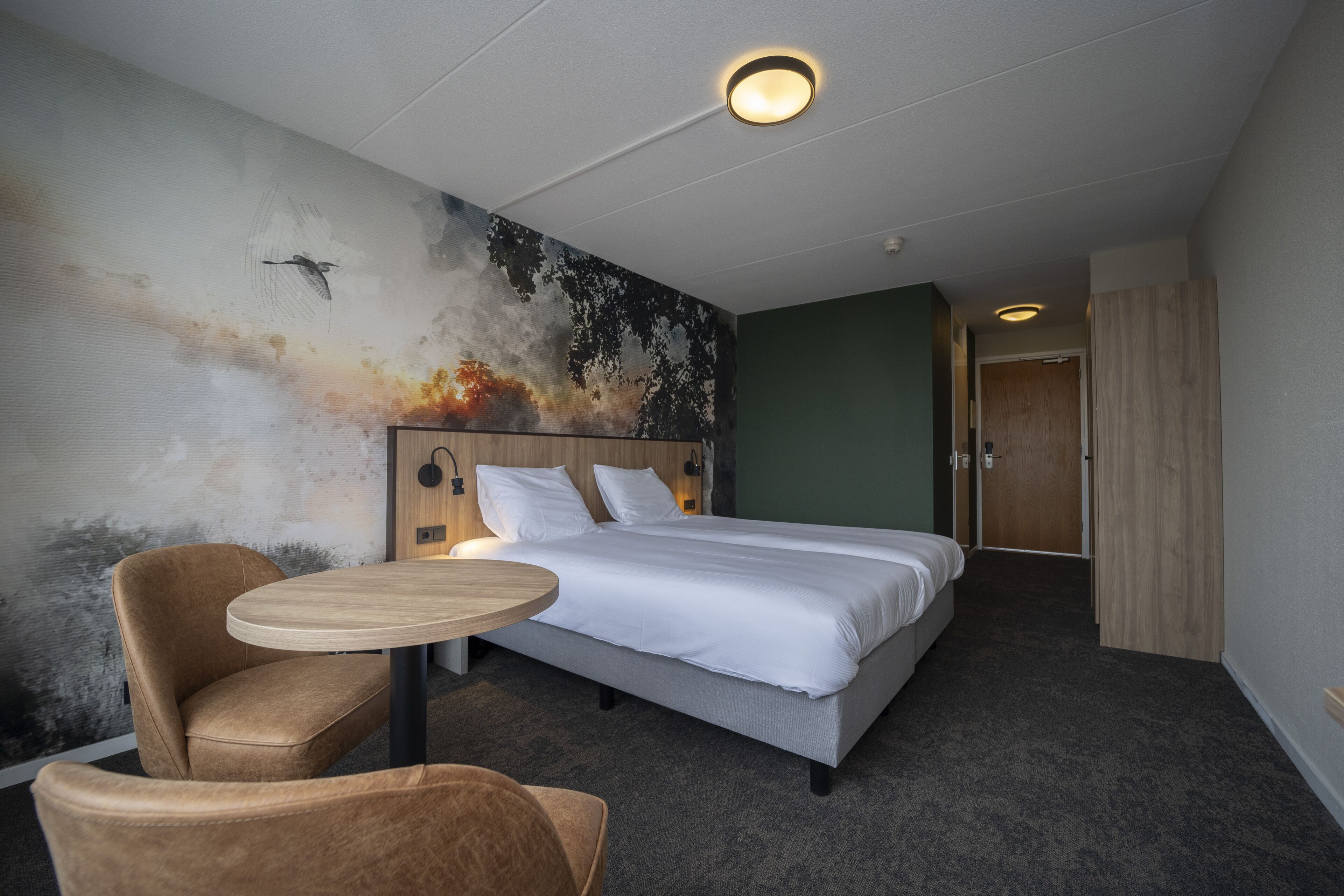 A hotel room with a bed, a round table, a chair, and a wall mural.