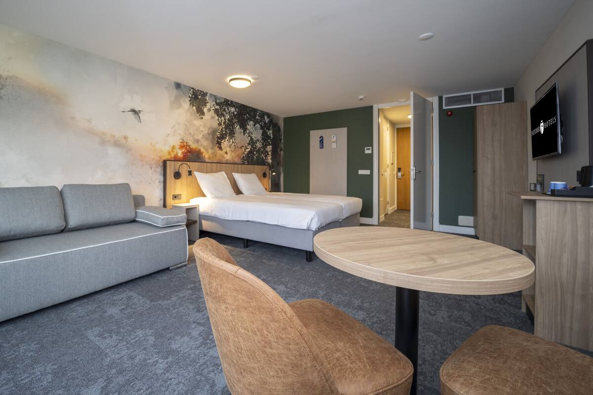 A modern hotel room with a grey sofa, a bed with white bedding, a round wooden table, and a TV mounted on the wall.