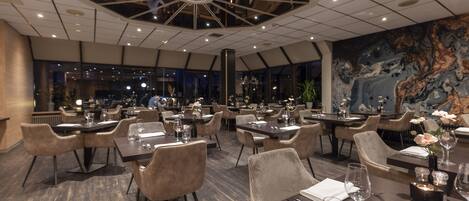A modern restaurant with multiple dining tables and chairs, a high ceiling with a geometric light fixture, and a large mural on the wall.
