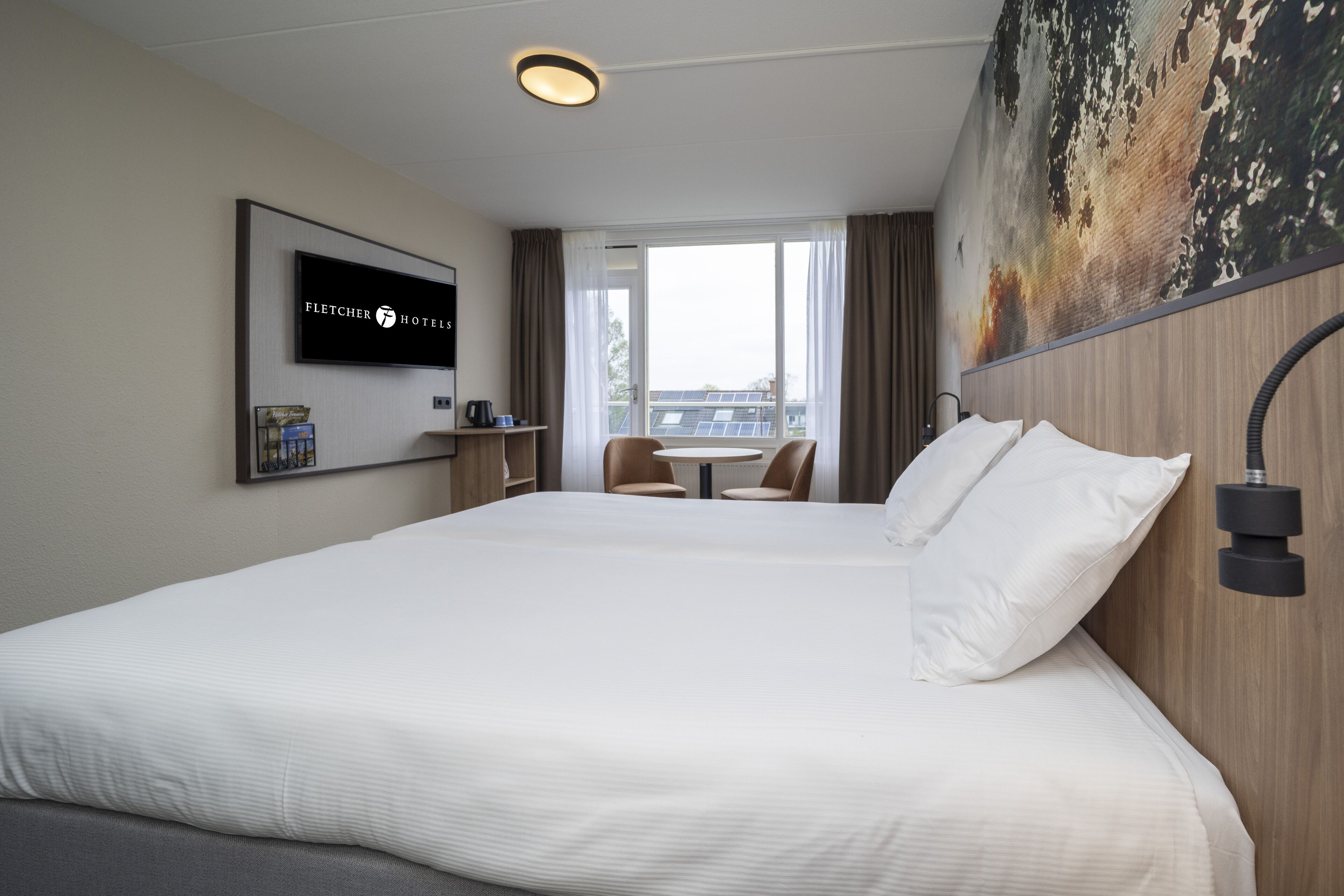 A hotel room with a large bed, a television mounted on the wall, a small table with chairs, and a view of the city through the window.