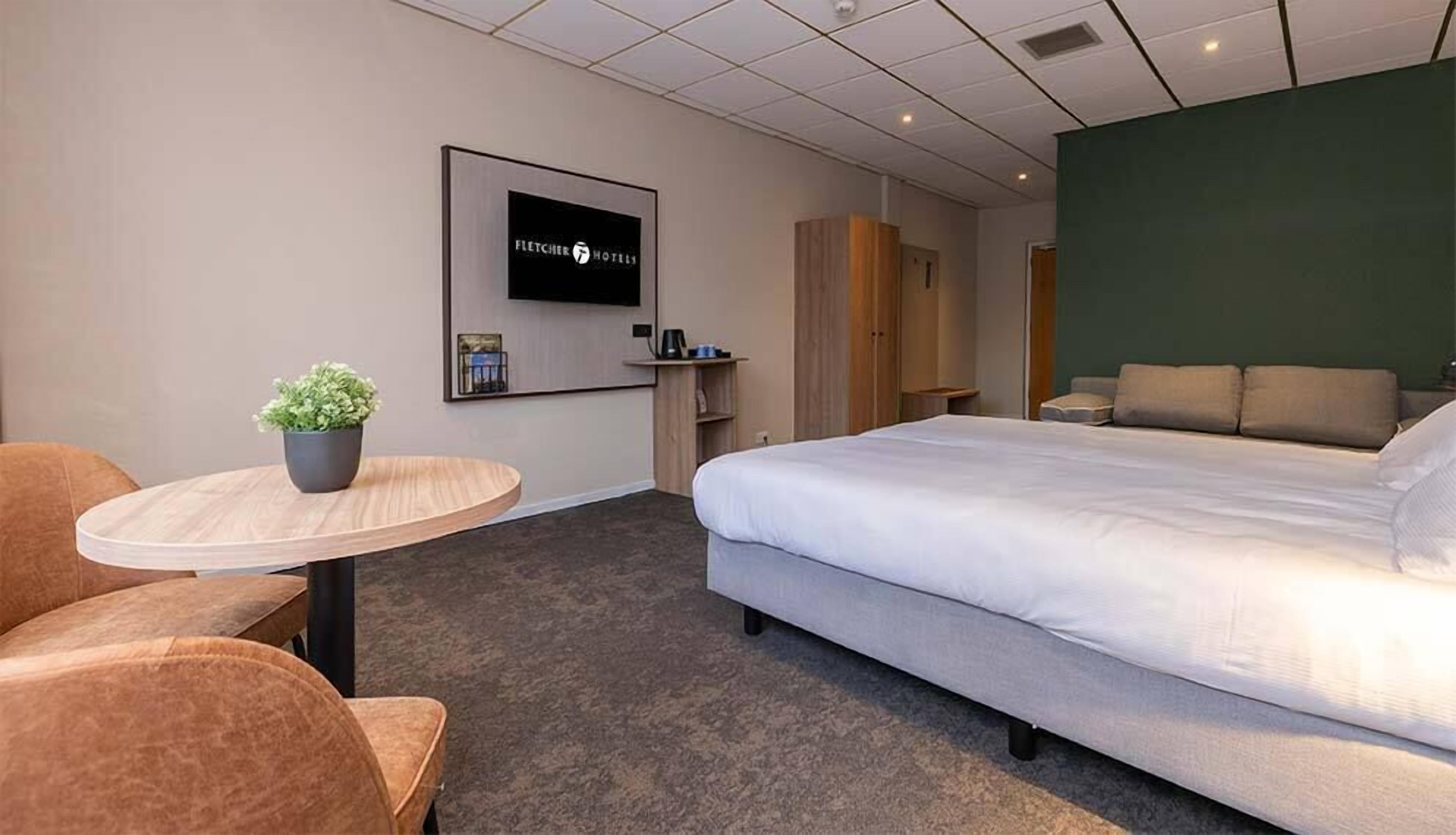 Quarto de hotel com uma cama, uma mesinha com uma planta, uma televisão fixada na parede e uma parede verde decorativa.