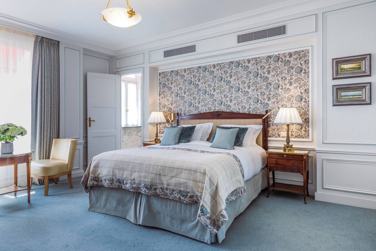 A bedroom with a large bed, two bedside tables with lamps, a chair, and a window with curtains.