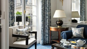 A room with a blue and white patterned sofa, a matching armchair, a small round table with a lamp, and a window with floral curtains. - Hôtel San Régis (Paris)