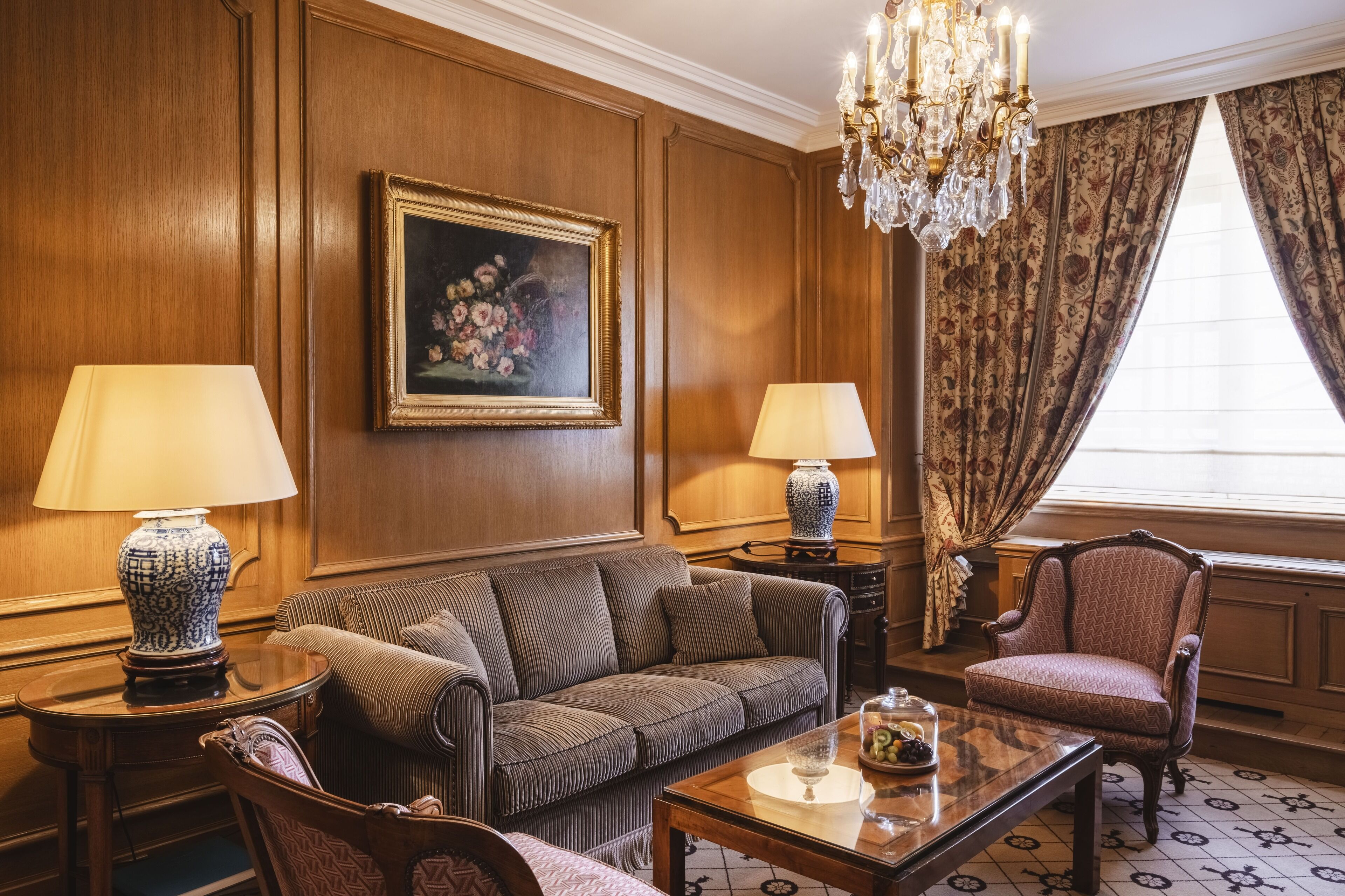 A room with a wooden paneled wall, a framed floral painting, a chandelier, a patterned sofa, a patterned armchair, two lamps, and a table with a glass jar.