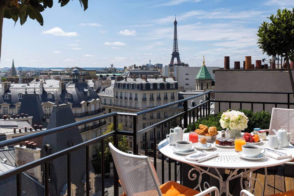 Un balcón con una mesa dispuesta para el desayuno, con vistas a un paisaje urbano donde se divisa la Torre Eiffel a lo lejos.