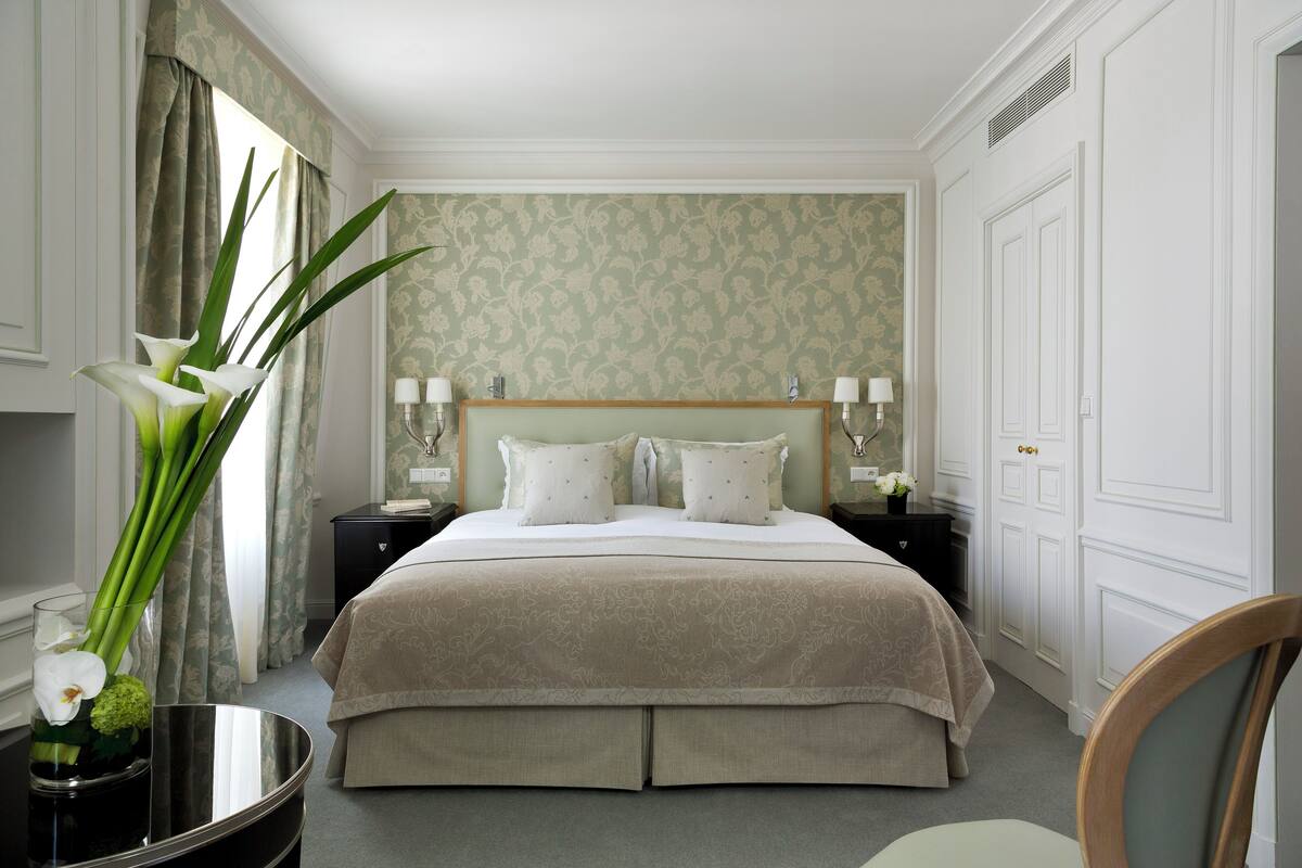 Habitación de hotel ordenada con una cama grande, una mesita de noche con lámparas, un florero con flores y una silla.