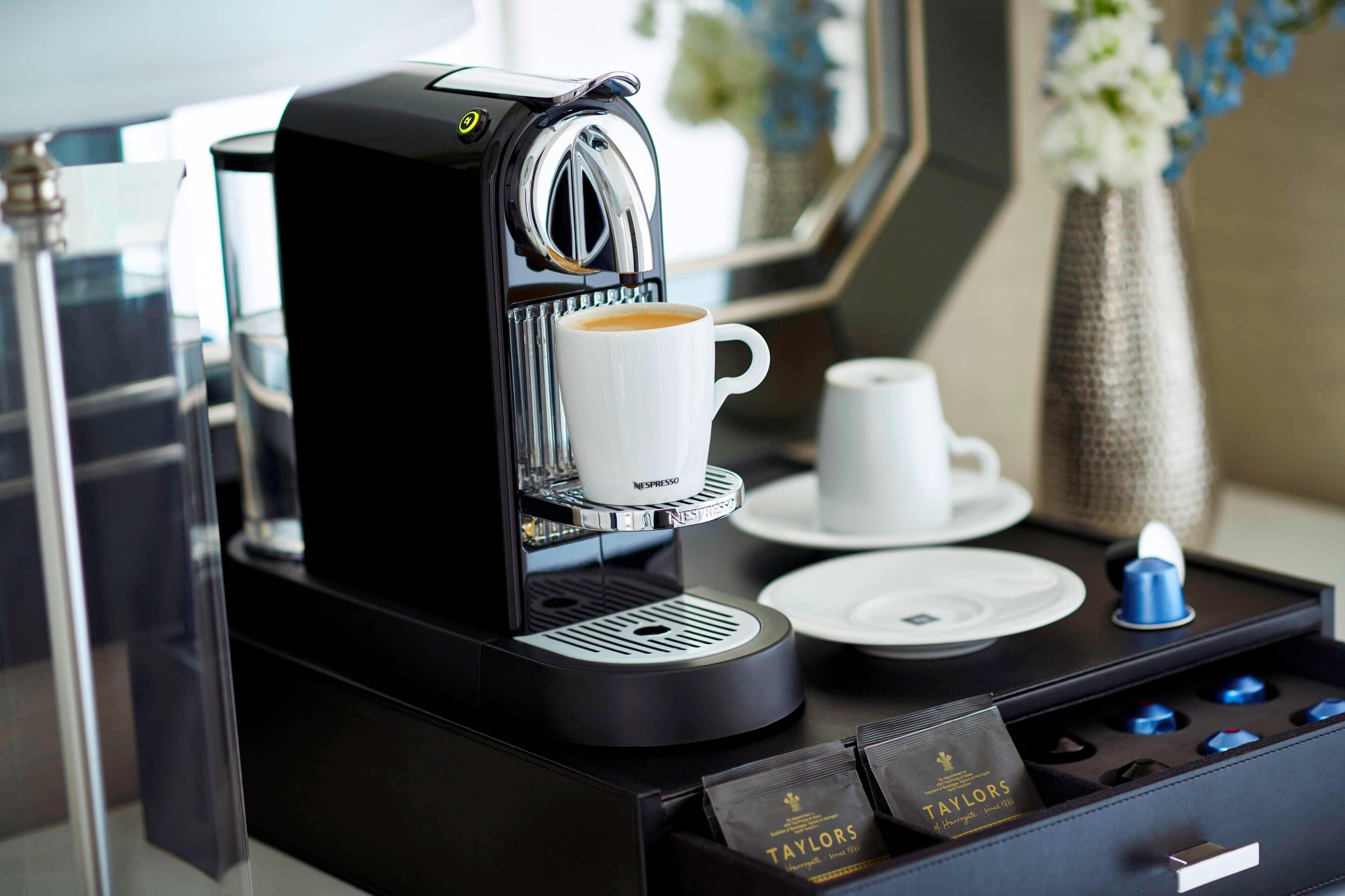 Uma máquina de café Nespresso com uma chávena de café, uma chávena branca, um prato e outra chávena azul numa bandeja.