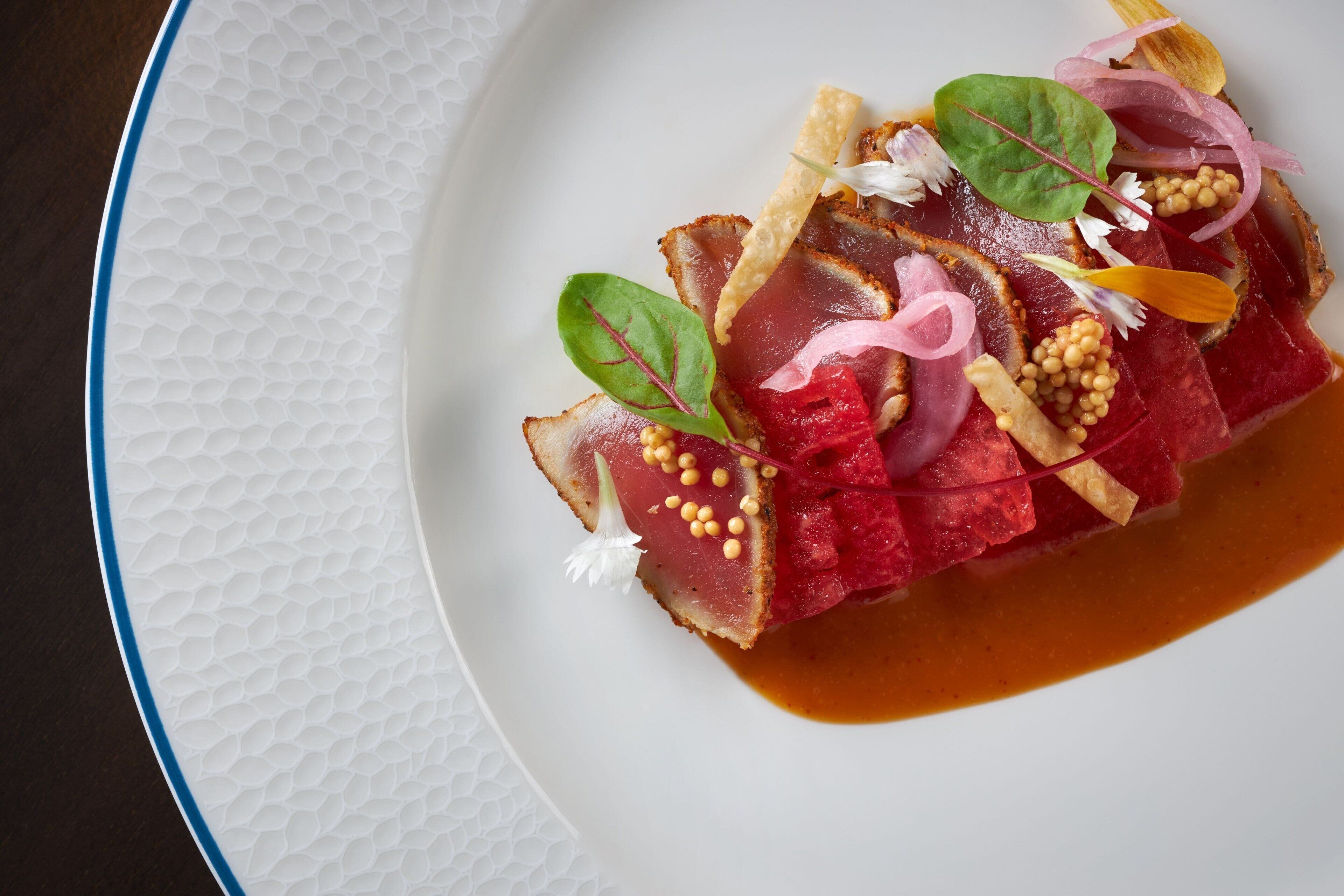 A white plate with a textured edge, containing a dish of thinly sliced meat, possibly tuna, garnished with greens, onions, and small round items, possibly seeds or nuts.
