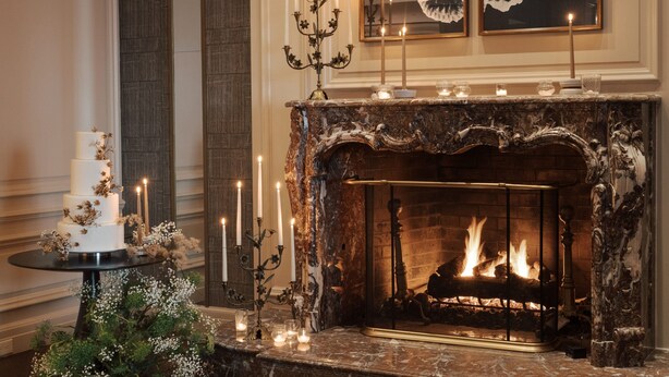 A grand fireplace with a fire, surrounded by candles and a decorative cake on a table.