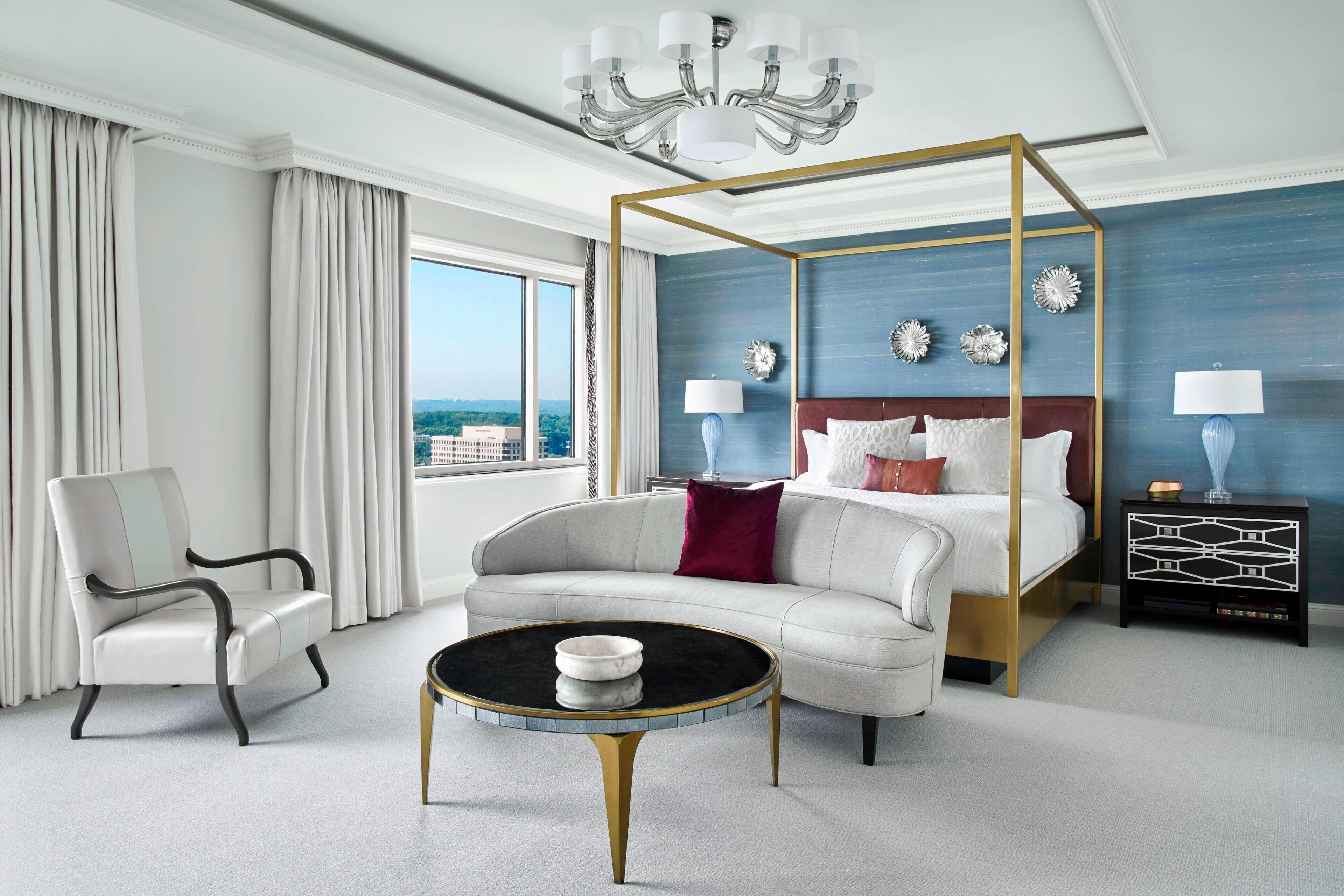 A modern hotel room with a four-poster bed, a round coffee table, a white sofa, and a view of the city.