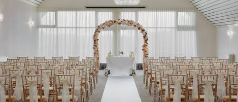 A wedding ceremony setup with rows of chairs, a white aisle, floral arch, and chandeliers.