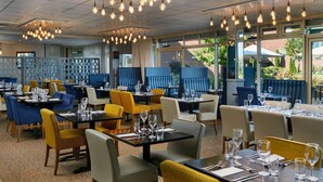 Un restaurante con varias mesas y sillas, un techo con luces colgantes y grandes ventanales que dan al exterior.